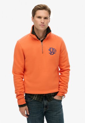 Oranje fleece trui met een halve ritssluiting en een geborduurd blauw logo. Draagt over een groen geruit overhemd. Heeft lange mouwen en een comfortabele pasvorm.