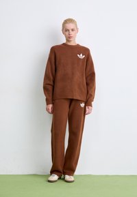 Ξανθός άνθρωπος που στέκεται σε πράσινο πάτωμα φορώντας καφέ πλεκτό πουλόβερ και παντελόνι, και τα δύο με λογότυπα Adidas σε λευκό, καθώς και μπεζ αθλητικά παπούτσια.