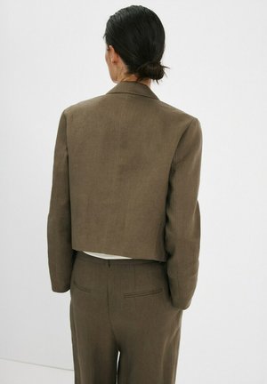 Persona con cabello oscuro recogido en un moño bajo, vistiendo un blazer corto verde oliva y pantalones a juego de talle alto, de espaldas sobre un fondo blanco.