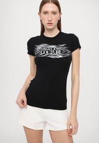 Must hõlmik must puuvillane T-särk lühikeste varrukatega ja ümmarguse kaelusega, millel on rinnal valge lainemustriga logo "VERSACE JEANS COUTURE".