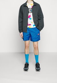 Sportjacket in donkergrijs, tie-dye T-shirt met meerkleurige vlekken, blauwe geblokte shorts, blauwe sokken en zwarte sneakers met witte accenten.