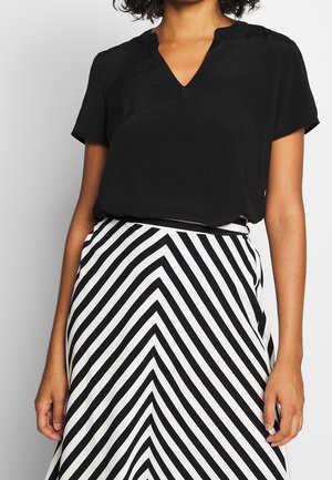 Persoon draagt een zwart V-hals T-shirt met korte mouwen, gecombineerd met een zwart-witte rok met chevron-patroon.