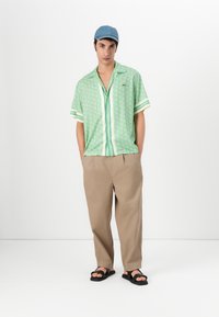 Green patterned short-sleeve shirt with a button-up front, paired with beige wide-leg pants and black flip-flops; denim cap completes the look.