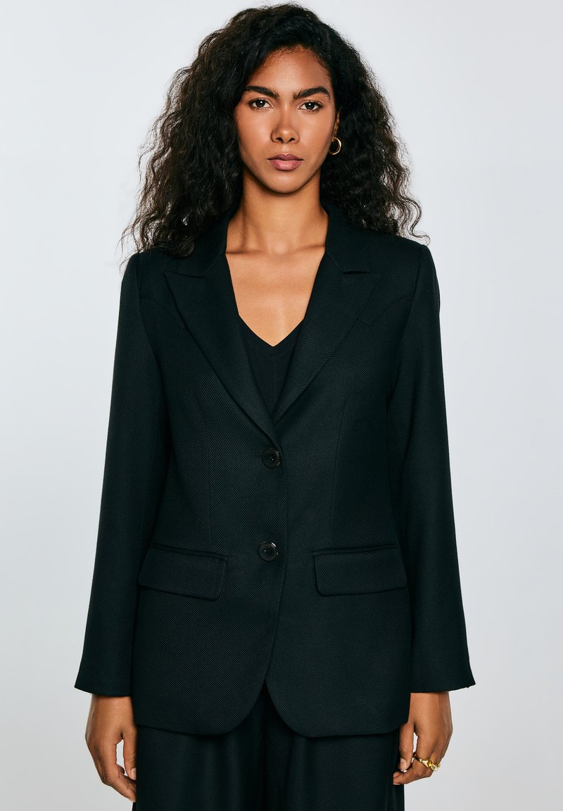 Blazer nero con vestibilità strutturata, rever a punta, due tasche frontali, chiusura con un singolo bottone e motivo a spina di pesce testurizzato.
