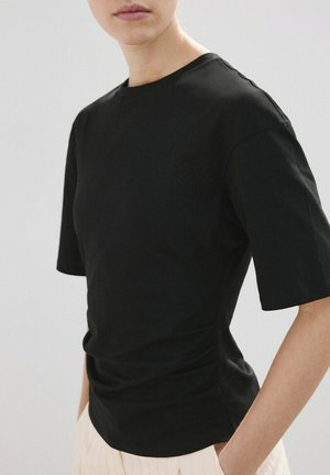 Ženska, oblečena v oprijeto črno majico s kratkimi rokavi, stoji ob navadni svetli podlagi z roko v žepu bež hlač.