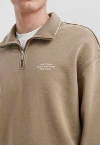 Beige sweatvest met rits, rib gebreide kraag en de tekst "KANT NIET OVER IETS MAKEN, GEWOON RELAXEN" gedrukt op de voorkant, gladde textuur.