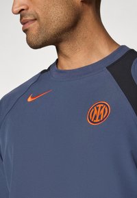Felpa sportiva blu con accenti neri, dotata di un logo Nike arancione e di un emblema circolare arancione. Tessuto morbido, scollo a girocollo.
