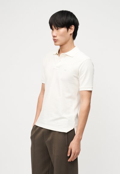 Νεαρός άνδρας που φοράει λευκό κοντομάνικο πόλο και καφέ παντελόνι, στέκεται με τα χέρια στις τσέπες μπροστά σε μια απλή σκηνή.