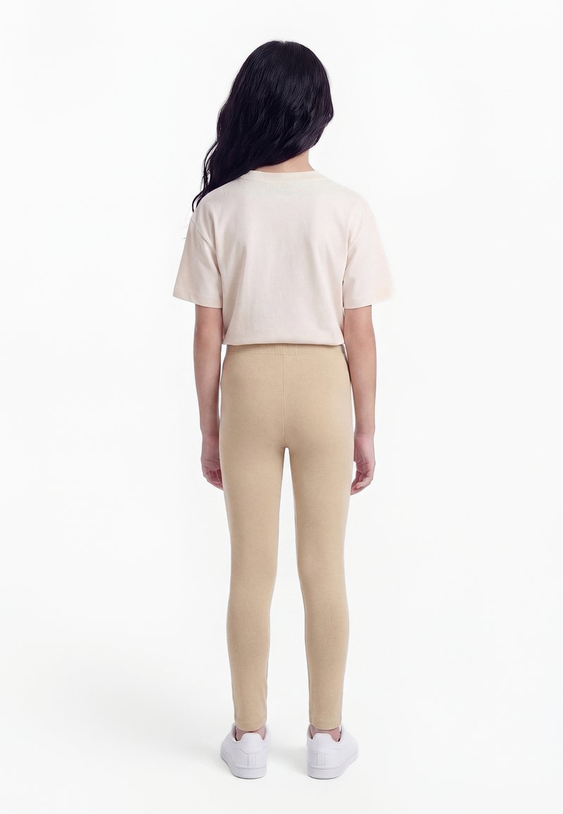Kind steht mit dem Rücken zur Kamera, trägt ein beigefarbenes Kurzarmhemd, beige Leggings und weiße Turnschuhe vor weißem Hintergrund.