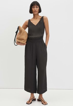 Frau trägt einen ärmellosen schwarzen Overall und schwarze Sandalen, hält eine beige geflochtene Tasche mit schwarzem Riemen, steht vor weißem Hintergrund.