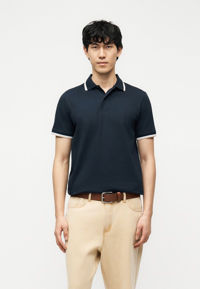 Jeune homme aux cheveux noirs portant un polo bleu marine avec des bordures blanches et un pantalon beige, debout devant un fond blanc uni.