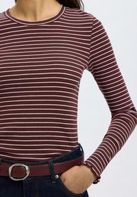 Burgunderfarget langermet topp med hvite horisontale striper, ribbet tekstur og rynkede mansjetter, kombinert med mørk denim og et matchende belte.
