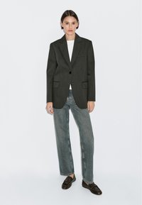 Blazer verde scuro, con texture, dotato di due tasche frontali e chiusura con un bottone, indossato sopra una maglietta bianca e abbinato a jeans larghi blu chiaro e scarpe scure.
