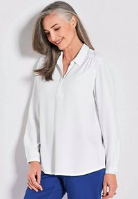 Weiße langärmlige Bluse mit Kragen und geknöpftem Verschluss; glatte Textur und seitliche Schlitze; kombiniert mit blauen Hosen.