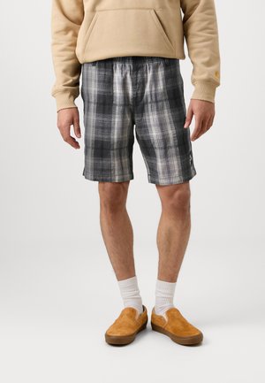 Άνδρας που φοράει γκρι καρό σορτσάκια, μπεζ φούτερ, λευκές κάλτσες και καφέ παντόφλες, στέκεται μπροστά σε απλό φόντο.