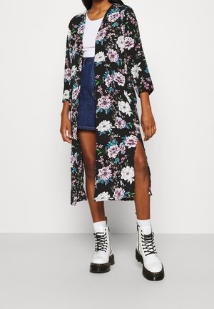 Vrouw draagt een zwarte, bloemetjes lange cardigan over een wit topje en een spijkerrok, gecombineerd met witte veterlaarzen en witte sokken.