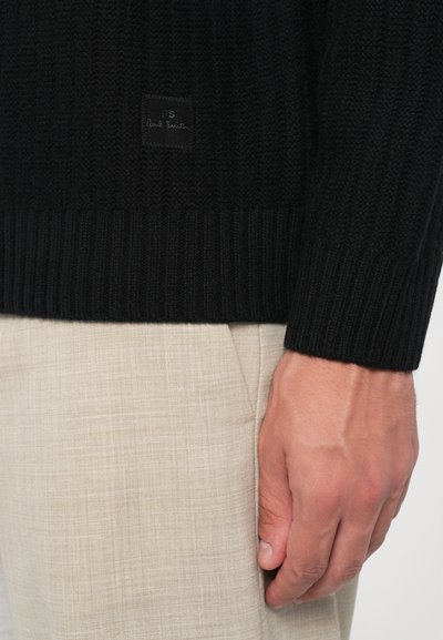 Prim-plan al unei mâini lângă pantaloni texturați bej purtați cu un pulover tricotat negru, ce prezintă o mică etichetă coasă.