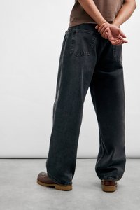 Svart vid bukse laget av myk denim, med synlige lommer og en løs passform, kombinert med brune sko mot en nøytral bakgrunn.
