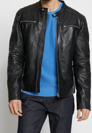 Homme portant une veste en cuir noire avec des poches zippées sur la poitrine par-dessus un pull en maille bleu et un jean foncé, torse visible.