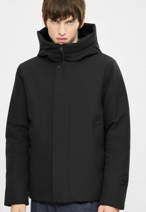 Young man wearing a black padded jacket with a high collar and zipper, standing against a plain white background.
