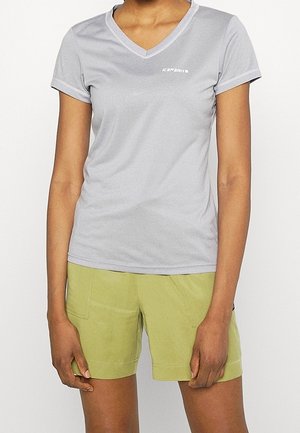 Femme portant un t-shirt gris clair à col en V et manches courtes et un short vert clair, debout les bras détendus le long du corps sur fond blanc.