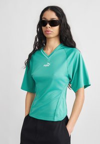 Γυναίκα που φοράει μαύρα γυαλιά ηλίου, μπλε-πράσινη κοντομάνικη μπλούζα Puma με V-λαιμόκοψη και μαύρο παντελόνι, με τα χέρια στις τσέπες, στέκεται μπροστά σε ένα απλό φόντο.