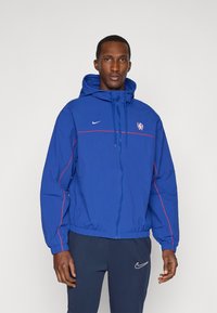 Blue Nike jacket with a hood, red accents, and zip closure. Features a logo on the chest and elastic cuffs and hem. Worn over navy pants.