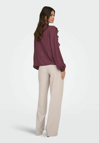 Blouse bordeaux avec des accents froncés, en tissu texturé, col rond et manches longues, associée à un pantalon large beige clair.