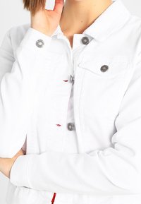 Biała dżinsowa kurtka z klasycznym kołnierzem, srebrnymi guzikami i dwoma przednimi kieszeniami. Gładka tekstura i dopasowany krój, odpowiednia na codzienne wyjścia.