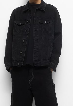 Person trägt schwarze Jeansjacke mit Knopfverschluss und schwarze Hosen mit sichtbaren Nähten, steht vor weißem Hintergrund.
