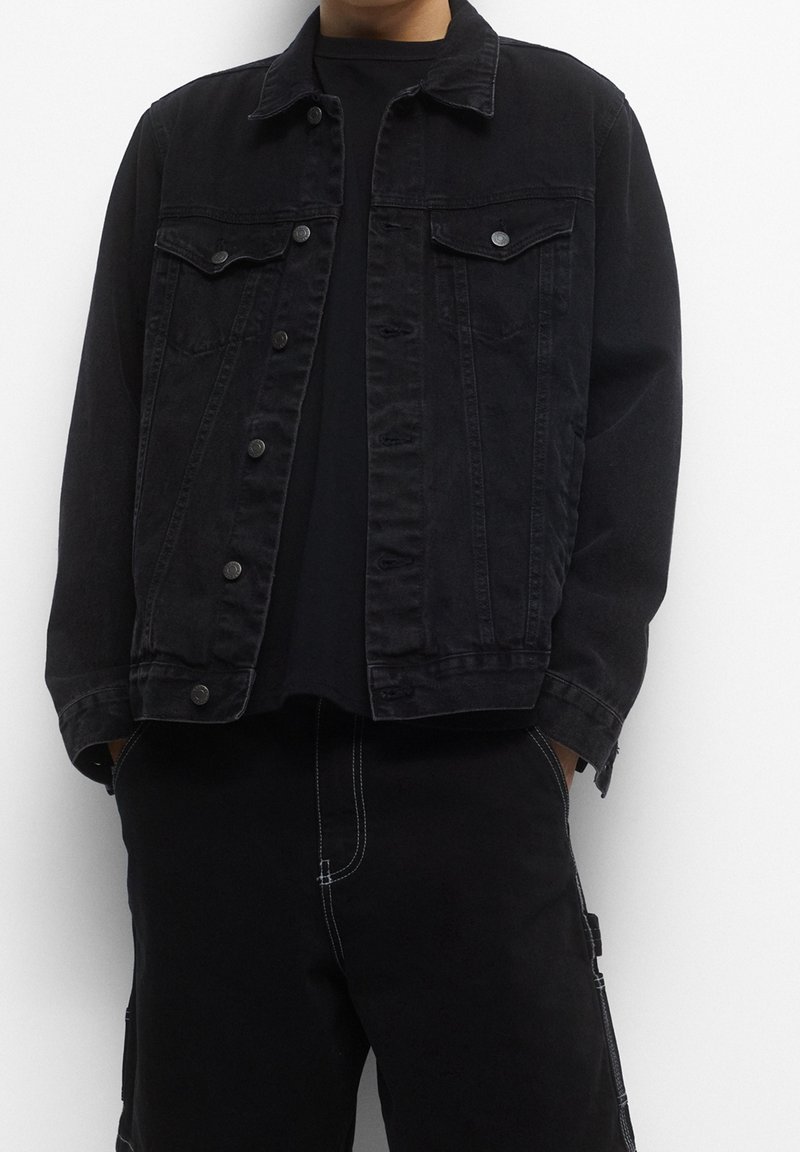 Personne portant une veste en jean noire avec des boutons et un pantalon noir avec des coutures visibles, debout devant un fond blanc.