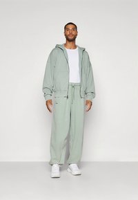 Šviesiai žalias per didelis džemperis su gobtuvu ir atitinkamos kelnės, pagamintos iš minkštos medžiagos, su priekinėmis kišenėmis ir laisvo kirpimo. Balti marškinėliai ir sportbačiai.