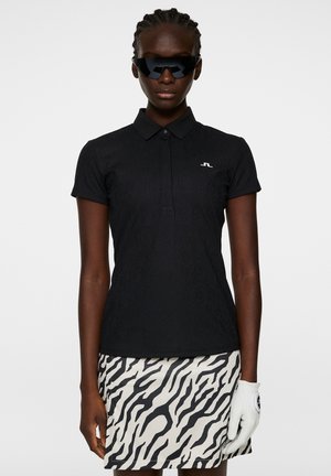 Schwarzes Poloshirt mit strukturiertem Finish und kurzen Ärmeln, kombiniert mit einem Rock im weißen und schwarzen Zebra-Design. Accessoires sind dunkle Sonnenbrillen und ein weißer Handschuh.