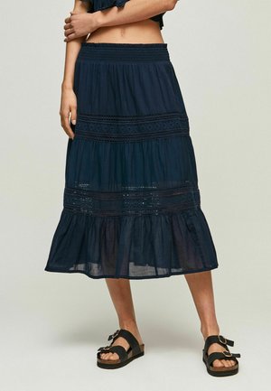 Femme portant une jupe midi bleu marine avec des détails en dentelle et des sandales noires avec de grandes boucles dorées, debout devant un fond uni.