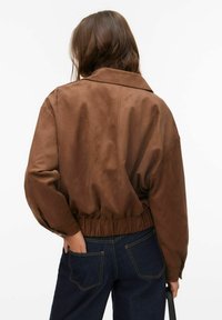 Smeđa, krojena jakna nalik brušenoj koži s klasičnim ovratnikom i elastičnim rubom, uparena s tamnim trapericama. Jednostavan dizajn, bez vidljivih metalnih dijelova.