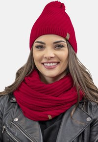 Rød strikket beanie med snoet top og matchende tekstureret infinity-scarf. Begge genstande har et diskret ribbet mønster og et lille mærke.