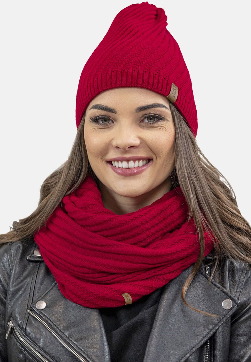 Rød strikket beanie med snoet top og matchende tekstureret infinity-scarf. Begge genstande har et diskret ribbet mønster og et lille mærke.