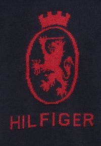 Tessuto a maglia blu navy con uno stemma di leone rosso ricamato all'interno di un contorno circolare, sormontato da una corona, e la scritta "HILFIGER" sotto.
