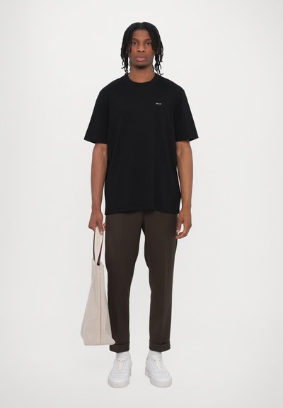 Un tricou negru supradimensionat cu un logo mic, asociat cu o pereche de pantaloni maronii și adidași albi. Persoana ține o geantă de pânză bej.