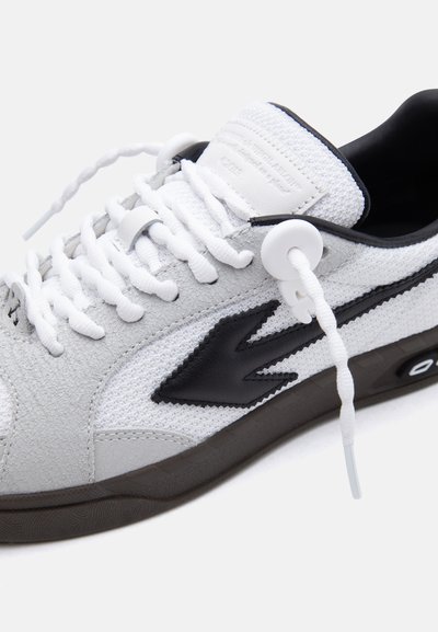Λευκό και μαύρο αθλητικό παπούτσι χαμηλού τύπου με πλεγμένα λευκά κορδόνια, μαύρο λογότυπο στο πλάι, υφασμένη επάνω επιφάνεια και μαύρη μαλακή σόλα.
