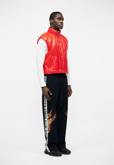 Άνδρας που στέκεται, φοράει λαμπερό κόκκινο αμάνικο γιλέκο κοντομάνικο φουσκωτό, λευκό μακρυμάνικο πουκάμισο, μαύρο παντελόνι με σχέδιο φλόγας και λευκή ρίγα στο πλάι με κείμενο, και μαύρα παπούτσια.