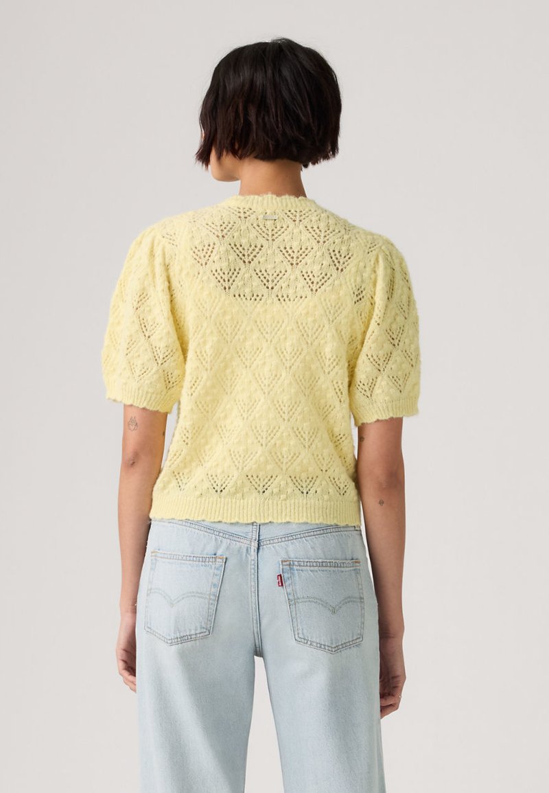 Person with short dark hair wearing a pale yellow knit short-sleeve sweater and light blue denim jeans, facing away.