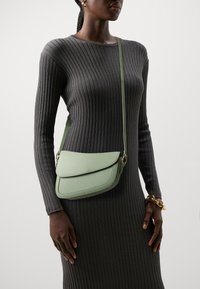 Femme à la peau foncée dans une robe grise à côtes portant un sac bandoulière vert clair et un bracelet en or chunky à son poignet droit.