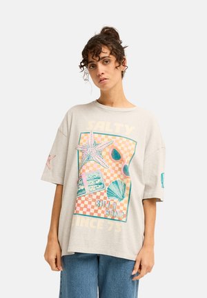 Jong persoon met krullend haar draagt een oversized beige T-shirt met zeesterren, zonnebril en schelpen, gecombineerd met een blauwe spijkerbroek.