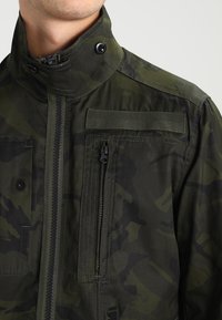 Man med en mörkgrön kamouflagejacka med hög krage, dragkedja framtill, knäppta flikar och en vertikal dragkedjeförsedd bröstficka.