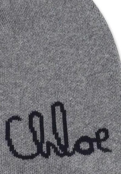 Tessuto a maglia grigio con testo in corsivo nero che scrive "Chloe" su una superficie tessile arrotondata, probabilmente un berretto o un cappello.
