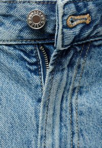 Primo piano di un tessuto denim blu chiaro che presenta un bottone in metallo, una cerniera e cuciture a contrasto, mettendo in risalto la texture e la trama del materiale.