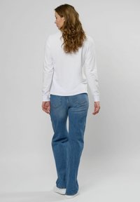 Sieviete ar viļņainiem brūniem matiem, tērpusies baltā krekliņā ar garām piedurknēm, zilās džinsās un baltās sporta kurpēs, stāvot ar muguru pret pelēku fonu.