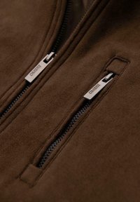 Brown suede jacket with two zippered pockets. Silver-tone zipper hardware features engraved brand name. Smooth texture throughout.