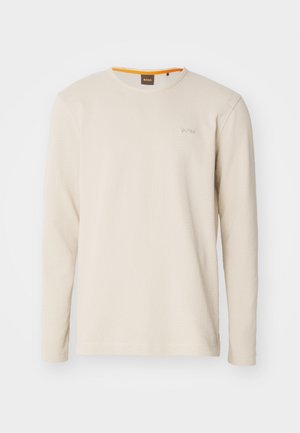 T-shirt beige clair à manches longues en tissu texturé. Présente un col ras du cou et un petit logo sur la poitrine avec un accent orange à l'intérieur du col.